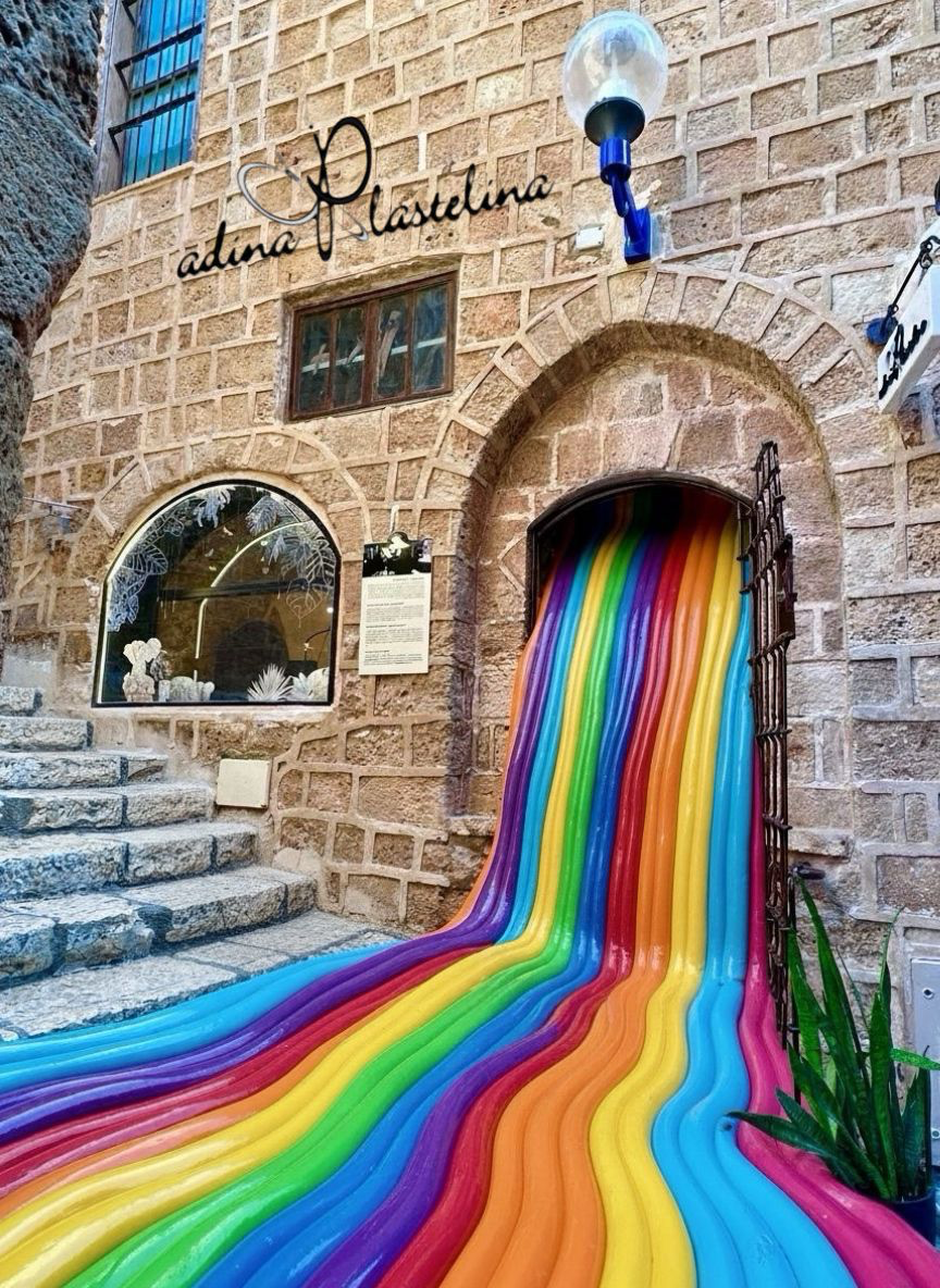 Photograph of the Adina Plastelina art installation in Old Jaffa showing the studio signage on the stone wall above a vibrant rainbow-colored flowing sculpture cascading down from an arched doorway, with decorative window displays and ancient Jerusalem stone architecture