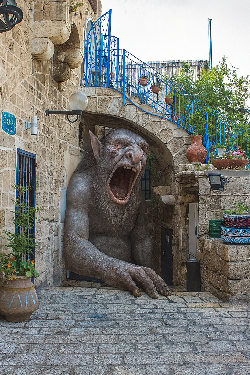 Dramatic fantasy composite of a massive Nordic troll creature emerging from a stone archway in Old Jaffa, with its mouth open in a roar, set against traditional architecture with blue railings and cobblestone streets